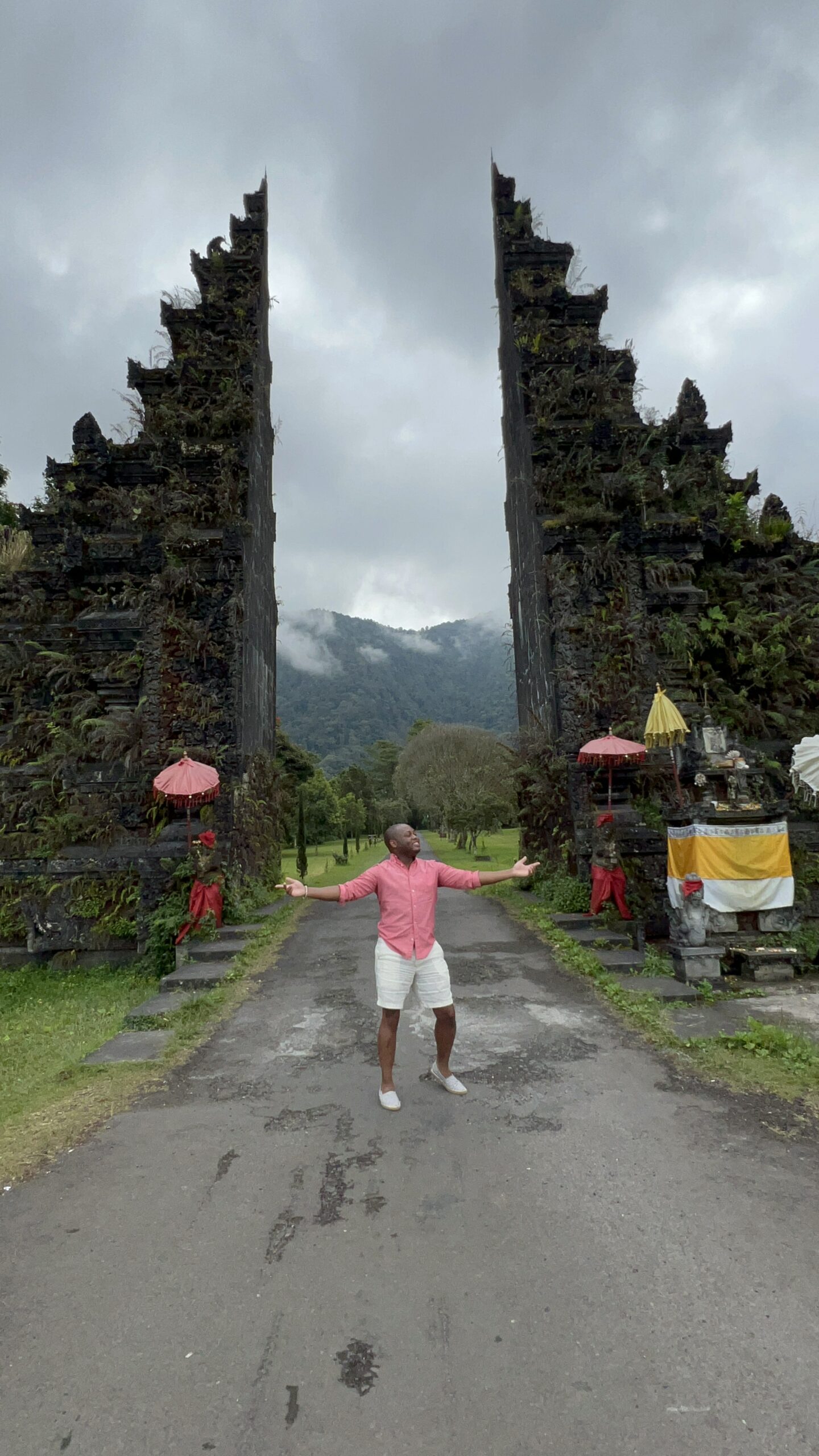 Man in a coral shirt and white shorts with arms spread wide, standing between the iconic split gates ('candi bentar') at the entrance to a Balinese temple, with misty mountains in the background