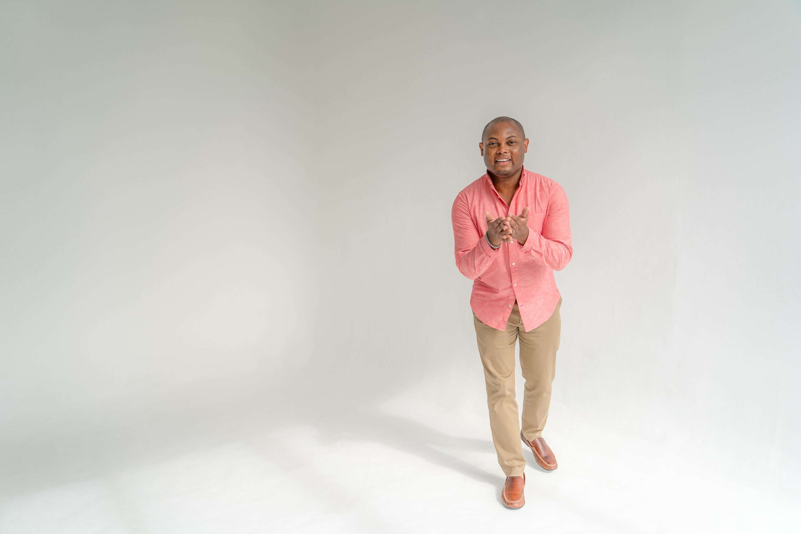 Confident man in a casual coral button-up shirt and khaki pants with brown shoes, standing with clasped hands and a pleasant expression in a bright, spacious studio