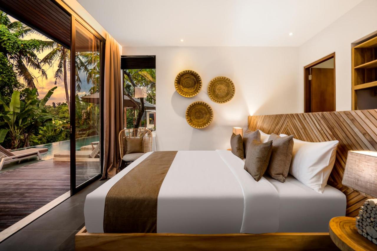 Elegant and modern bedroom with a comfortable bed with white linens and brown accent pillows, wooden headboard, decorative golden wall plates, floor-to-ceiling glass door opening to a tropical view with lush greenery, and natural light enhancing the serene ambiance.