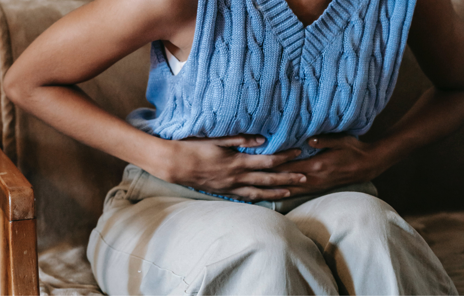 Close-up of an unidentifiable individual seated in a brown vintage armchair, wearing a sleeveless blue cable-knit sweater and beige trousers, with hands clasped over the abdomen, possibly suggesting rest or discomfort, in a room with soft, natural lighting.